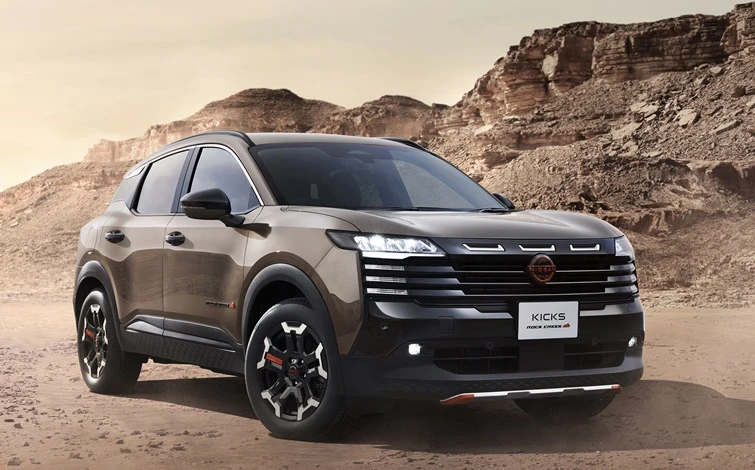 A Kicks ROCK CREEK in a beige-toned body color is parked in a front three-quarter view facing right. The black-accented front grille, unique alloy wheels, and Lava Red accent parts create a rugged and powerful appearance. In the background, barren rocky mountains and cliffs with layered rock formations stretch across the landscape, while dry terrain covered with sand and small stones extends into the distance. Illuminated by natural light, the vehicle's tough SUV styling stands out in this off-road environment.