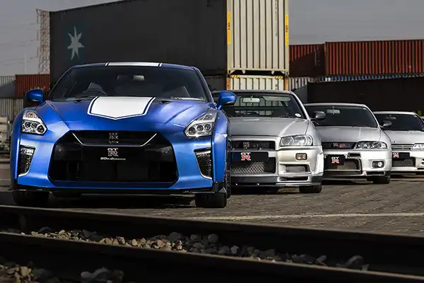 Multilple of silver racing cars with a blue racing car in the front parked neatly in front of shipping containers.