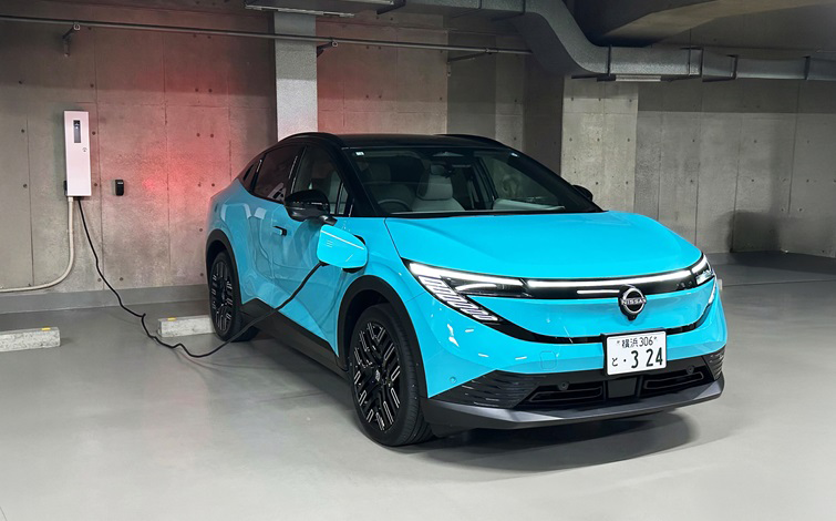 地下駐車場で、ターコイズブルーの車体の日産リーフが左前方から見える向きで駐車し、左側面から延びたケーブルを壁の充電器につないで充電している。