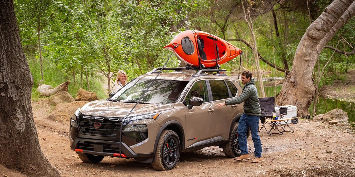Front left 3/4 angle of a 2026 Nissan Rogue parked on a camp site with a kayak on its roof and a man opening the door.