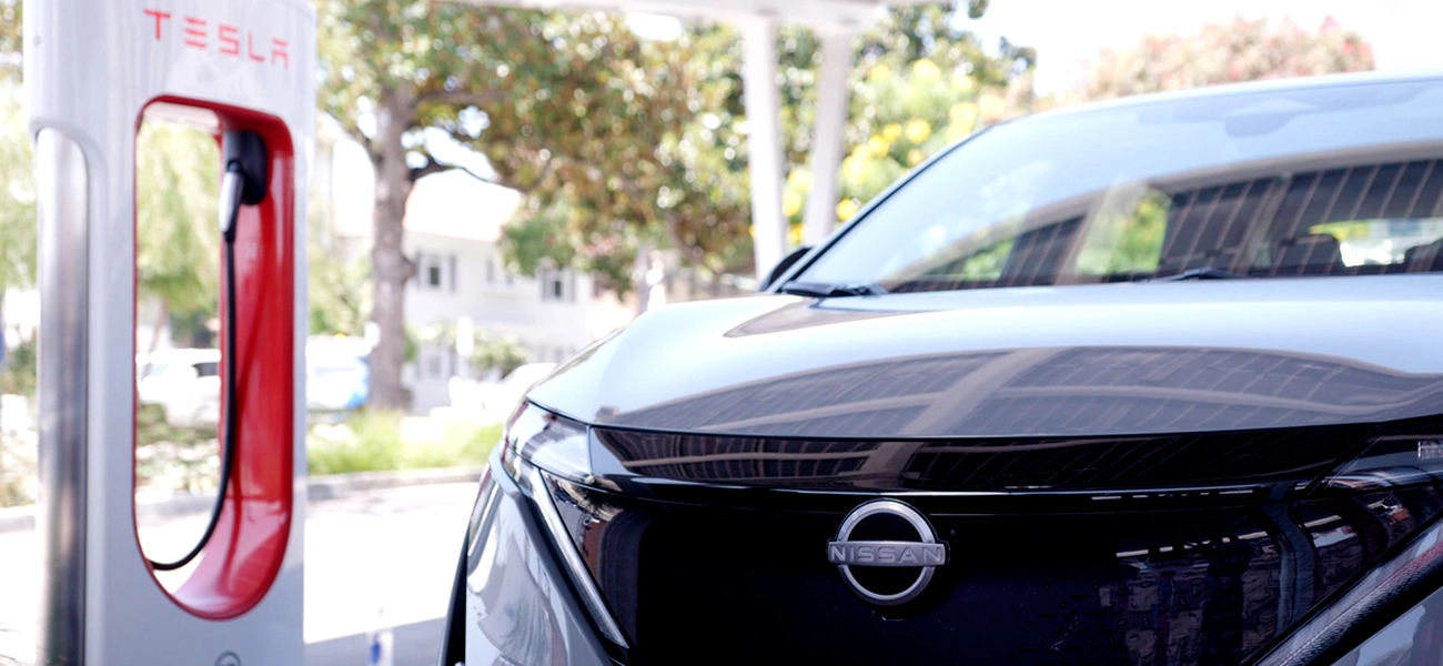 A front view of a gray 2025 Nissan Ariya parked by a charging station on a sunny day.