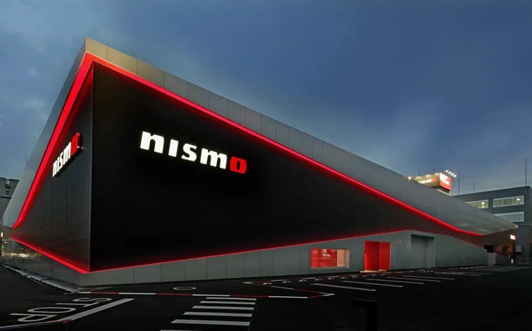 A NISMO showroom building with black and silver exterior walls is angled to the right against a dusk sky. Red LED lights outline the structure, and the front displays the 'nismo' logo in white and red. In the foreground, there is a black asphalt parking area with white road markings.