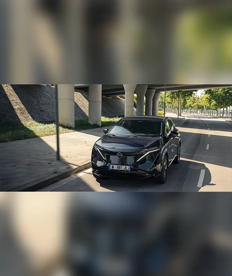 A black Nissan Ariya solar-powered concept car drives along a quiet, tree-lined city road beneath an overpass.