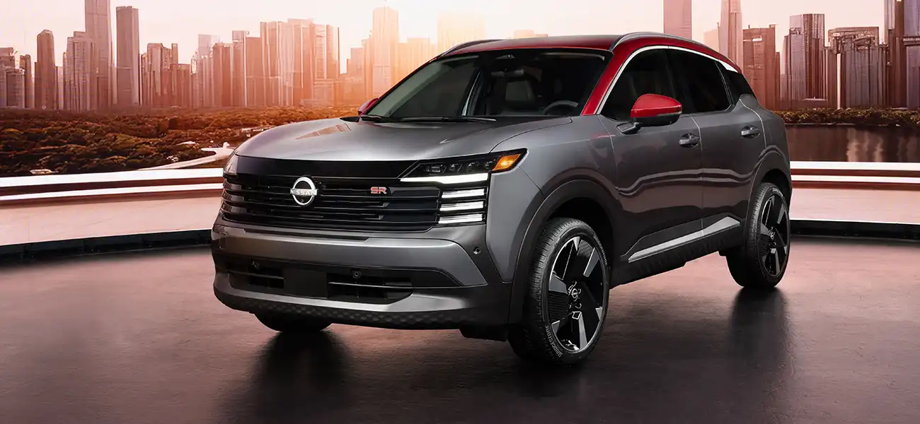 Front left 3/4 shot of a silver 2025 Nissan Kicks parked on concrete against a cityscape.