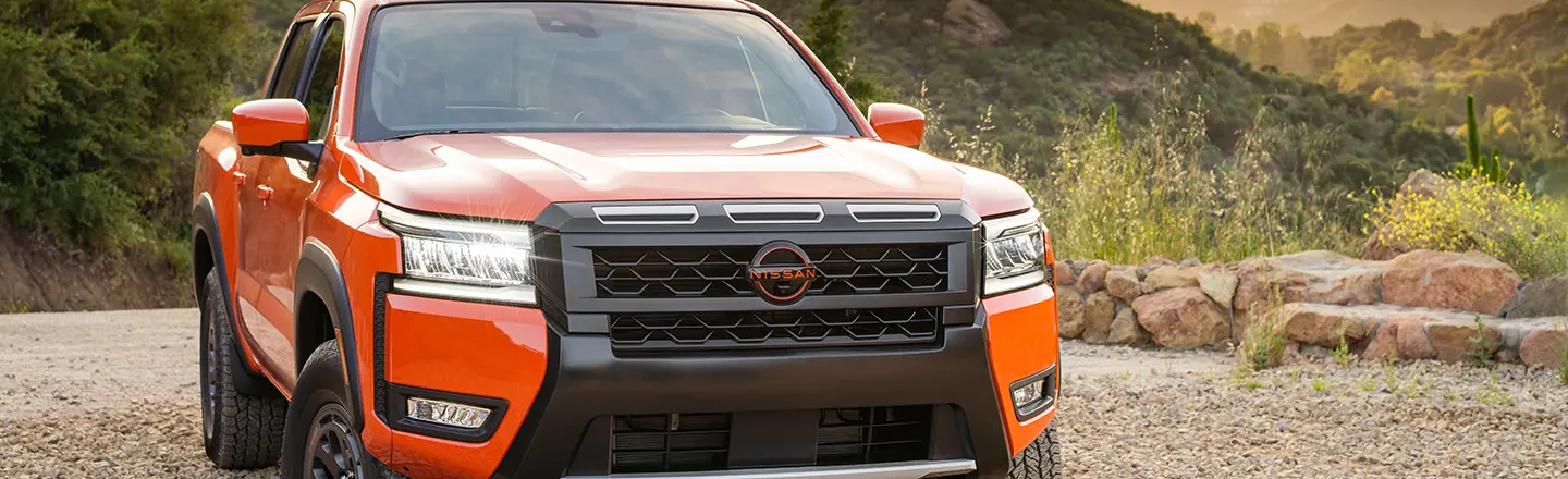Front right 3/4 angle of a red 2025 Nissan Frontier parked on gravel with tree covered mountains behind.