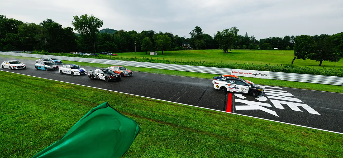 Multiple Sentra Cup racecars racing on a black track, bright green grass is beside and a green flag is in view.