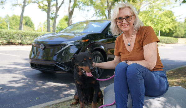 Henriëtte James and her rescue dog, Tula, smile as they sit in front of their black Nissan Aria parked on a leafy road.