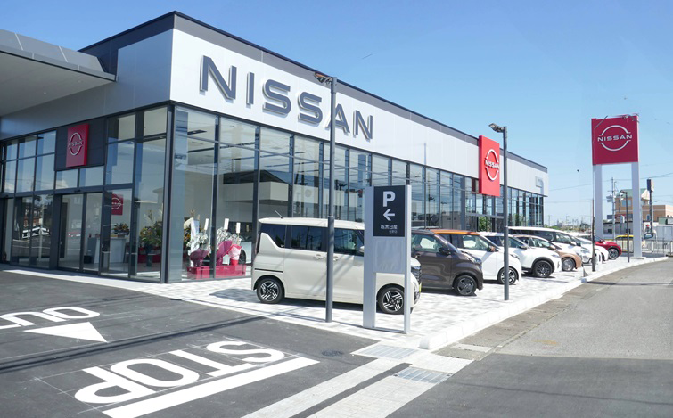 栃木日産佐野店の外観。ガラス張りのショールームとNISSANのロゴ看板、駐車場には複数の車両が並んでいる晴天の店舗風景。