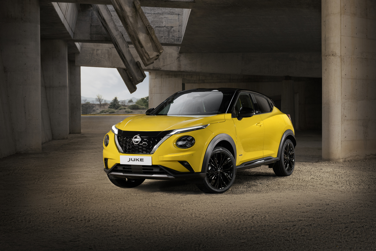Żółty Nissan Juke stojący przodem pod kątem na tle ściany, z drzewami w oddali