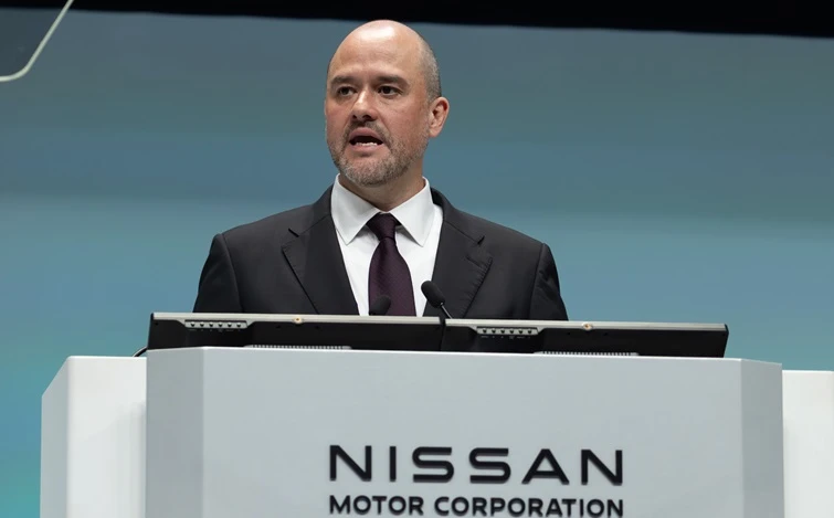 日産自動車のロゴが入った白い演台の前で、スーツ姿の人物がやや左を向いてスピーチしている様子。背景は青緑色のシンプルな壁面で、会見や発表の場面とみられる。