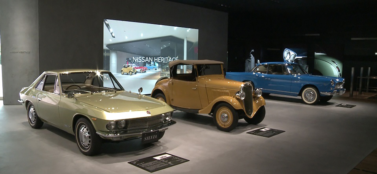 3 autos patrimoniales estacionados frente a una pantalla que dice "Nissan Heritage".