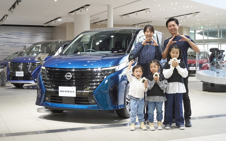 青い日産セレナがショールーム内に展示されている。車両は正面を向いており、特徴的なフロントグリルとヘッドライトが際立っている。背景には白を基調とした明るい室内空間が広がり、奥には同じモデルの車両が複数並んでいる。車両の前には5人の家族連れ（夫婦、子供3人）が立っており、車と一緒に両手で三角形の「おにぎり」ポーズを取ったり、実際におにぎりを持っている。